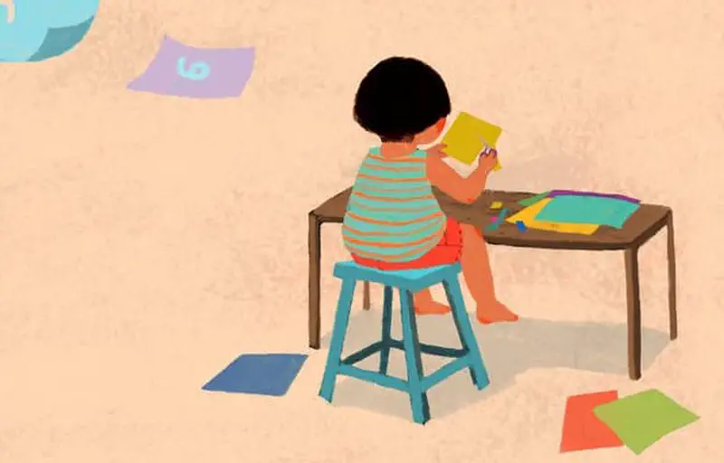 Boy seen from behind sitting at table drawing and cutting colorful paper crafts