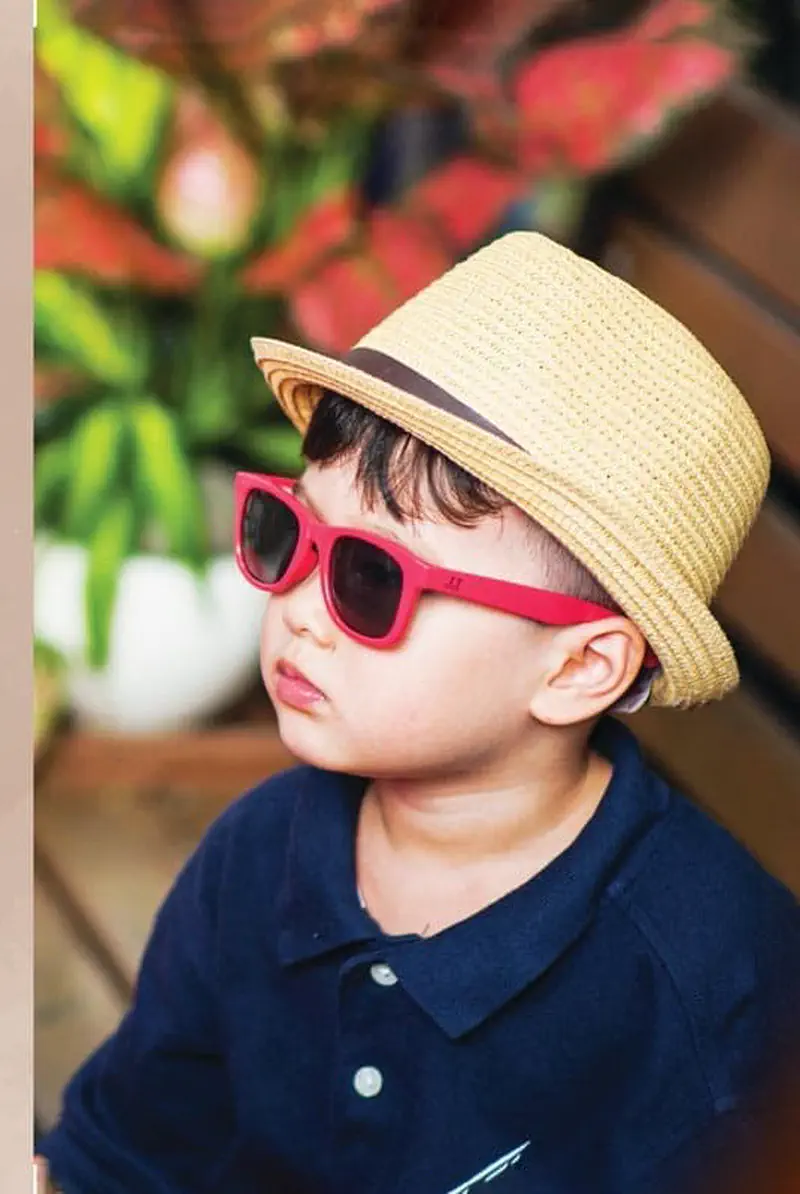 A young boy wearing a straw fedora hat with a brown band and red sunglasses, dressed in a navy polo shirt.
