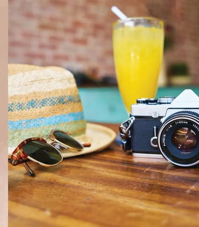A striped straw hat with blue and yellow bands sits on a wooden table next to sunglasses, a camera, and orange juice.