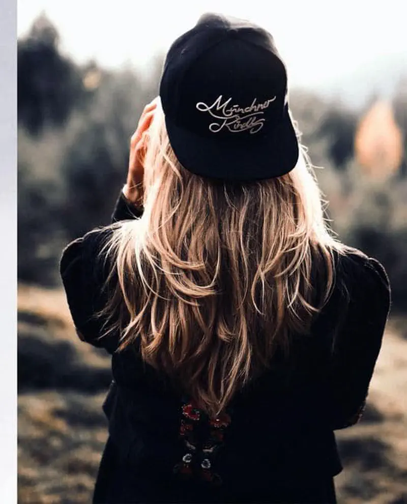 Back view of a woman with long blonde hair wearing a black baseball cap with cursive text, looking at a misty landscape.