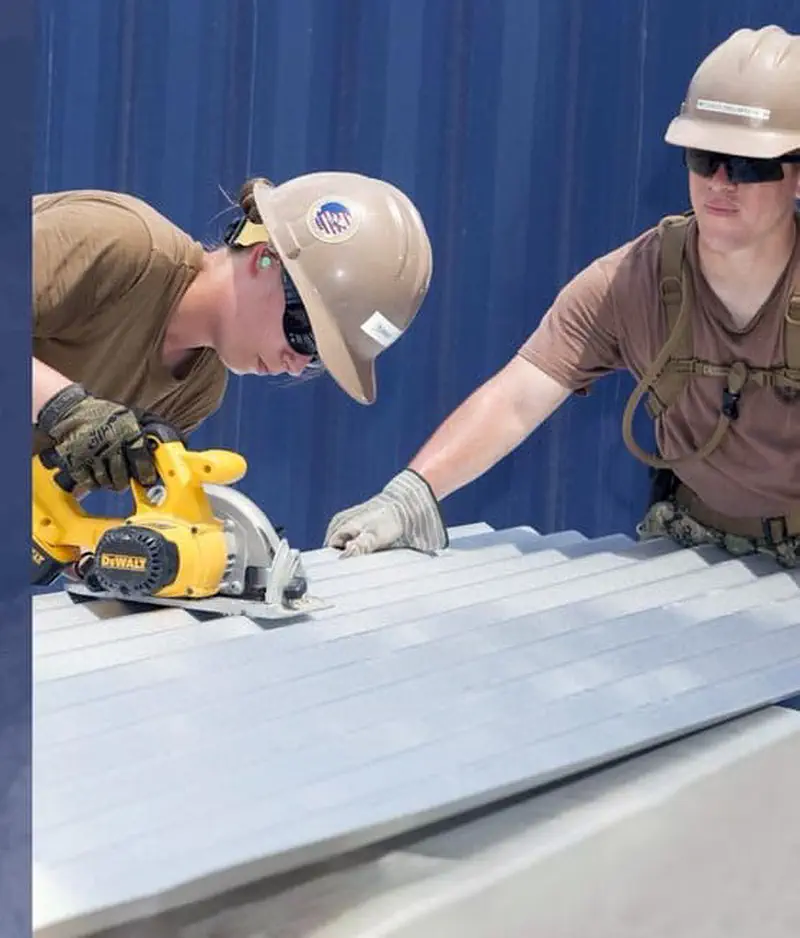 Two construction workers wearing tan hard hats while cutting corrugated metal roofing with a circular saw.