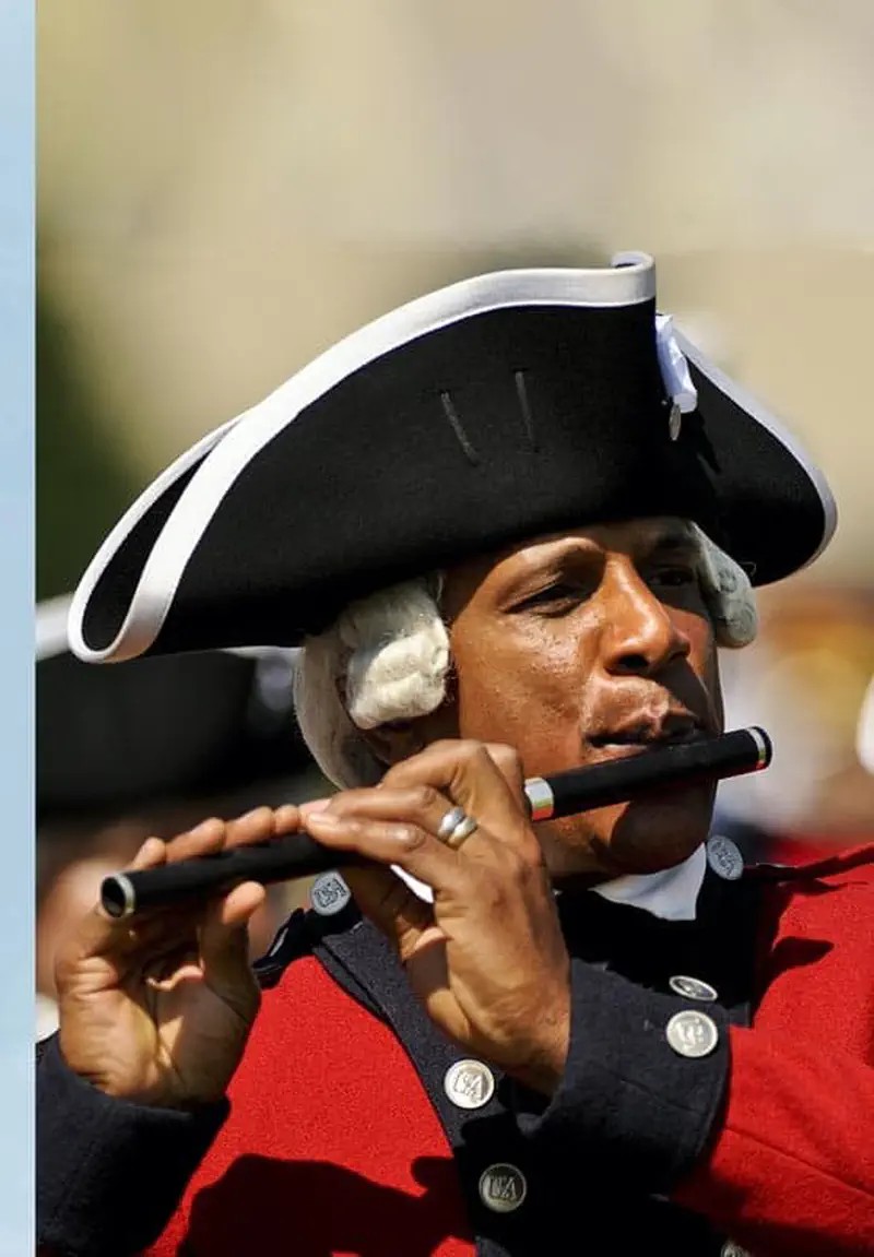 A man in colonial costume playing a fife while wearing a black tricorn hat with white trim and a grey wig.