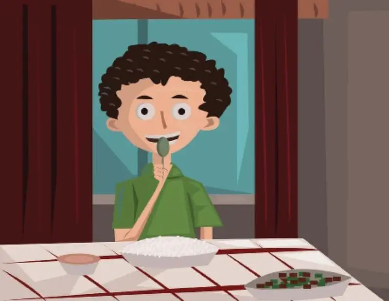 A boy with curly hair in a green shirt eating a meal with rice and vegetables at a table near a window with red curtains.