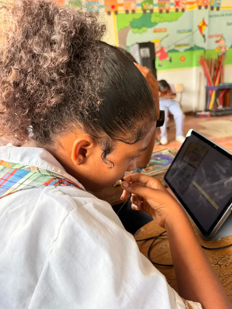 A student in Colombia holds the headset microphone near their mouth while reading a Spanish book aloud to Bookbot on a tablet, with a colourful alphabet mural on the classroom wall behind