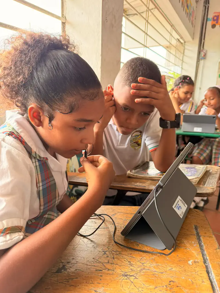 Two students in a Colombian classroom read with Bookbot on a tablet during the pre-pilot, one holding the headset microphone close while reading aloud and the other listening closely